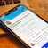 A smartphone displaying a social media profile is placed on a wooden table next to a laptop. The profile includes a picture, name, and details about a creator and photographer. The blue and white interface is visible on the phone screen.