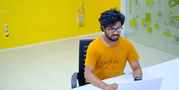 A person with glasses and a beard is sitting at a white desk, working on a laptop. They are wearing an orange t-shirt with text on it. The surroundings include a brightly colored office space with a predominantly yellow theme. There is a wall with the words 'Digital Agency' and a glass partition featuring playful icons.