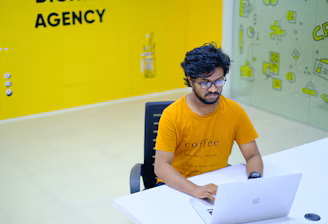 A person with glasses and a beard is sitting at a white desk, working on a laptop. They are wearing an orange t-shirt with text on it. The surroundings include a brightly colored office space with a predominantly yellow theme. There is a wall with the words 'Digital Agency' and a glass partition featuring playful icons.